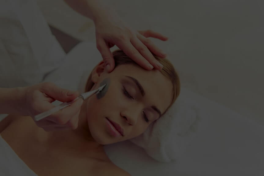 Person lying down with eyes closed, receiving a facial treatment with a brush applying cream.